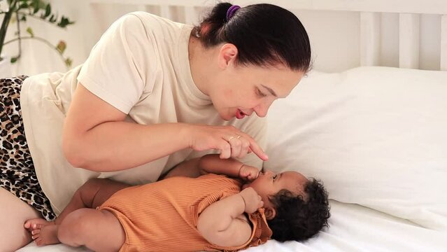 white Caucasian mom hugs and kisses an African-American black foster baby girl at home on the bed, maternal love and care, a mother of European appearance with a black child in her arms