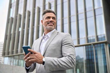 Happy business man investor of middle age in suit holding smartphone, mature professional businessman leader using mobile phone looking away standing in busy office city street with cellphone in hand. - 777387592