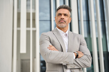 Confident middle aged business man wearing suit looking away outside office. Serious rich mature businessman investor wearing suit standing arms crossed looking away thinking or future success. - 777390324