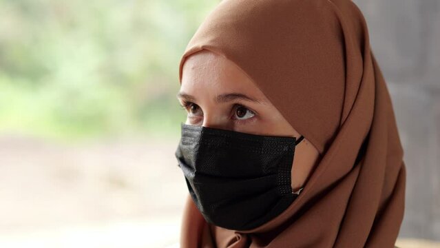 Young woman wearing black face mask and hijab, raise eyes and look up aside. Portrait shot of one person following health control measures during pandemic time