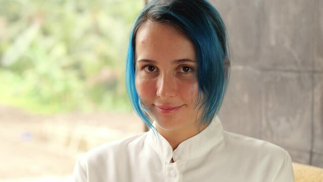 Positive attitude from young woman, she look straight to camera. Her hair is dyed to blue color, portrait shot with blurred background. Friendly smile on the face, she move head slightly