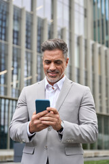 Happy middle aged business man ceo entrepreneur standing outside office using mobile cell phone. Mature businessman professional executive investor holding smartphone working on cellphone. Vertical. - 777403140