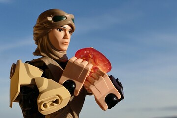 Naklejka premium LEGO Star Wars large action figure of Rey Skywalker in desert scavenger outfit, carrying half sliced cherry tomato, blue skies with scattered clouds in background, morning spring sunshine. 
