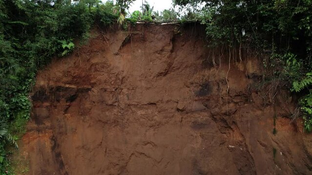 Top edge of recent landslide, exposed sandy earth, damaged road, tropical thickets around. Aerial camera fly up, showing open cavity at steep slope of ravine. Local calamity caused by heavy rainfalls