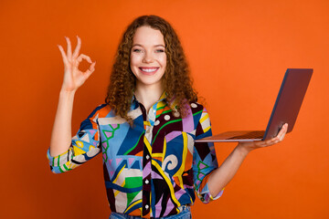Photo of charming adorable girl wear stylish print clothes hold netbook device okey gesture isolated on vivid orange color background