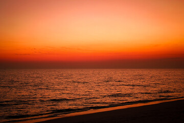 The sea after sunset has an orange glow and an orange sky with no clouds.