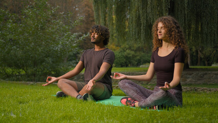 Two diverse multiracial guy and girl meditating zen meditation morning sport practice in park lotus position on yoga mat on grass sporty Indian Arabian man Caucasian woman relaxing in nature outdoors