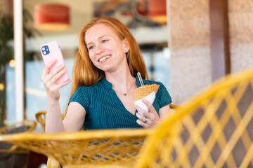 Happy redhead woman looking her phone on a terrace eating an ice cream
