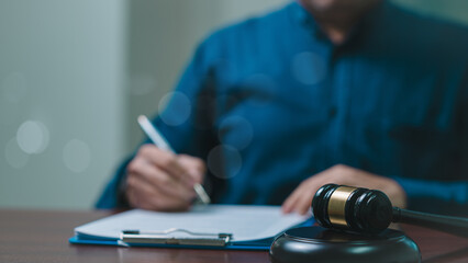 A lawyer man is writing on a piece of paper in front of a gavel. The gavel is on a desk and the man...