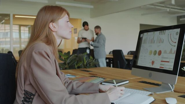 Businesswoman analyzing financial data on computer and writing down notes in copybook while working at desk in open space office