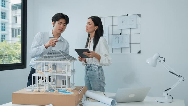 Beautiful young caucasian project manager brief asian engineer by using tablet while engineer measuring house model on meeting table with blueprint, architectural equipment scatter around. Immaculate