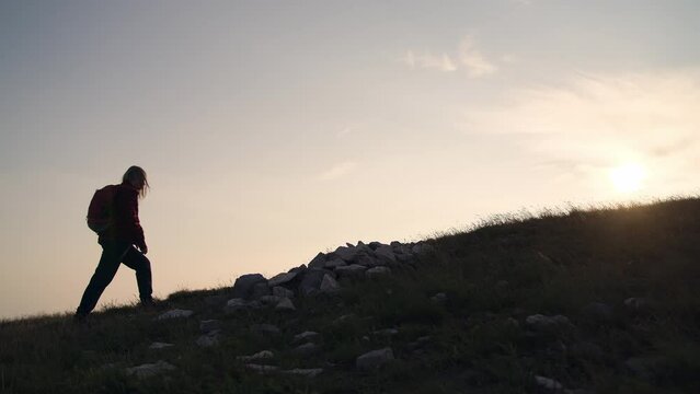 Female hiker silhouette with a backpack walking and following the trail uphill, enjoying the sky at sunset, low angle wide shot. Inspiration and healthy lifestyles concepts.