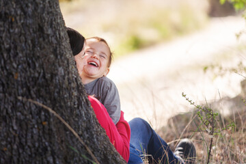 Mother and young son sitting behind the trunk of an oak tree on a spring day and laughing.