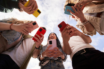 Low angle view of group happy unrecognizable people in circle using colorful mobiles phones together outdoor. Young friends and smiling Asian woman in center holding cells. Gen z technology addiction