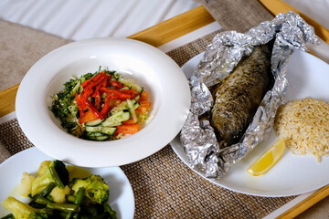 Lunch with fish, salad, rice and boiled vegetables and lemon on wooden tray in hotel room. Tasty nutrition, resting and relaxing in cozy hotel. Healthy meal for lunch or dinner.