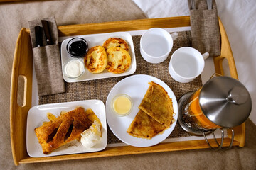 Condensed milk, sour cream and jam, breakfast cutlery. Wooden tray with food pancakes, cheesecakes, pastry with tea and ice-cream in hotel room. Tasty nutrition, resting and relaxing in cozy hotel.