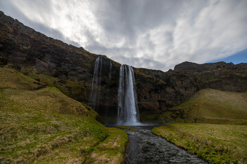 Fototapeta premium Skogafoss waterfall cascades down surrounded by mountains and green grass on a cloudy day, creating a picturesque natural landscape of fluvial landforms. Iceland