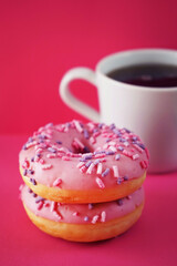 Donuts in pink glaze with sprinkles next to a white cup of coffee on a pink background