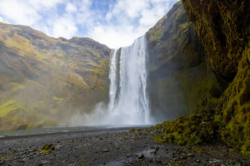 A stunning Skogafoss waterfall cascades down a mountain chute in a valley, surrounded by towering...