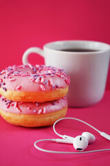 Donuts in pink glaze with sprinkles next to a white cup of coffee and headphones laid out in the shape of a heart on a pink background