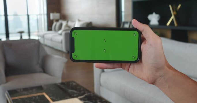 Young man hand holding mobile phone with horizontal green screen phone at living room. Male hand using mobile phone with green screen chroma key at home. Browsing internet, watching video content