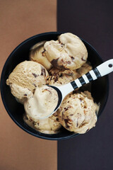 Nut ice cream balls with chocolate in a black bowl and a black and white spoon on a brown background