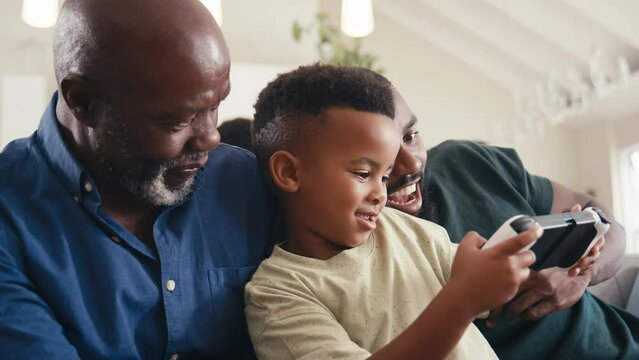 Grandfather with father and grandson playing handheld computer game at home with family in background - shot in slow motion