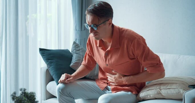 Man sits on couch at home and suffers from severe stomach pain. Hand of a mature guy suffering from pain, diarrhea or indigestion holds his stomach. Guy presses on his stomach because of pain