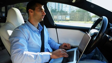 Handsome entrepreneur working on notebook while riding an autonomous self driving electric car at urban road. Male businessman typing text on laptop during riding on electrical vehicle with autopilot