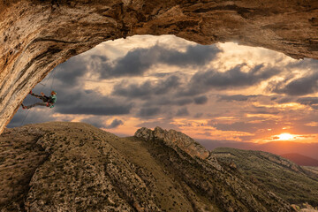 A man is climbing a mountain with a sunset in the background. The sky is cloudy and the sun is...