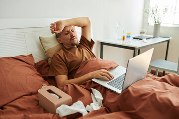 Sick man spending day in bed, sneezing and watching videos on laptop