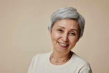 Cheerful mature woman with short grey hair and natural makeup looking at camera with smile during photo session on beige background