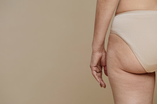Hand and buttock of senior female model wearing beige underwear standing in isolation against pastel background during photo shooting