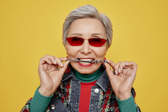 Aged woman in sunglasses and dopamine dressing biting her necklace with healthy teeth while posing in isolation on yellow background