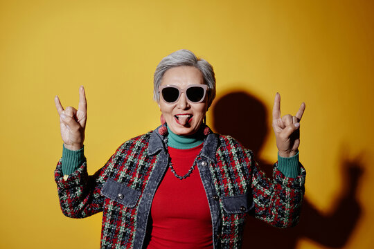 Positive senior woman in sunglasses and dopamine dressing showing her tongue during photo session against yellow background