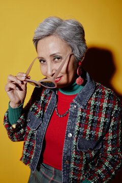 Flirty senior woman with short grey hair taking off sunglasses and looking at camera while posing in isolation against yellow background