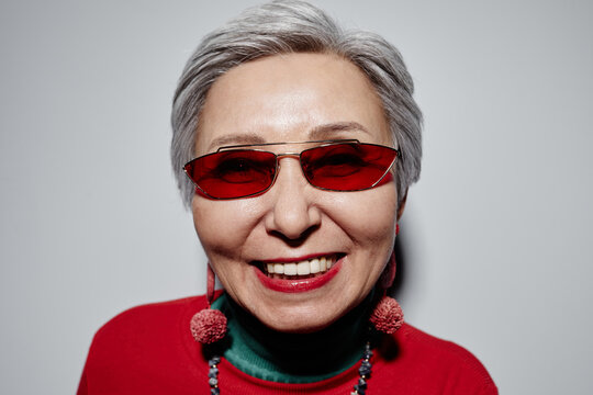 Face of cheerful senior woman in stylish sunglasses looking at camera with toothy smile against white background during photo session