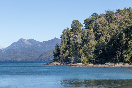 Horizontal photo of mountains full of vegetation and rocks, surrounded by tranquil waves of a lake's water, with a clear blue sky during a sunny day, in Lago Nahuel Huapi, Argentina