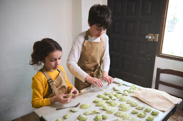 Fototapeta premium Adorable kids in the rural kitchen, rolling out dough with a wine bottle, sculpting dumplings with mashed potatoes filling. Cooking homemade vegetarian dumplings, Italian ravioli or Ukrainian varenyky