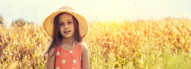 Happy little girl in a straw hat walks through a wheat field on a sunny summer day. Horizontal...