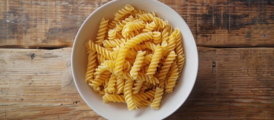 Overhead view of a white bowl filled with fusilli pasta on a wooden table. - obrazy, fototapety, plakaty