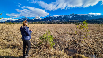 Woman holding pussy willows in spring with scenic view of majestic snow capped mountain peak Mittagskogel in Karawanks, Carinthia, Austria. Landscape of Finkensteiner Moor covered by dry golden grass - obrazy, fototapety, plakaty