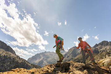 Two people are climbing a mountain together. One of them is wearing a backpack. The sun is shining...