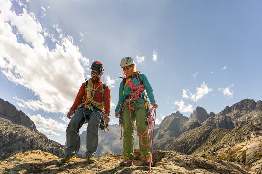 Two people are standing on a rocky mountain, one of them wearing a red jacket. They are both wearing harnesses and are looking at the camera
