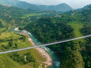 Aerial view of a Tibetan suspended bridge in Nepal is a primitive type of bridge in which the deck lies on two parallel load-bearing cables that are anchored at either end. Wild nature - 777643352