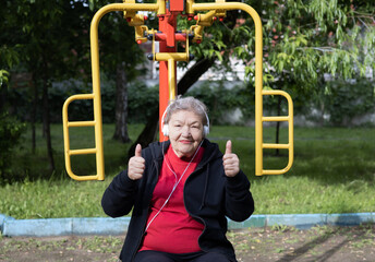 Smiling elderly woman in headphones is doing exercises on simulator, trainer outdoors in the yard. Active life of pensioners. Adaptation of pensioners in the modern world. Prevention of mental illness