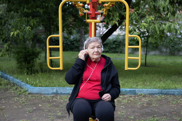 Smiling elderly woman in headphones is doing exercises on simulator, trainer outdoors in the yard and listening music. Active life of pensioners. Prevention of mental illness
