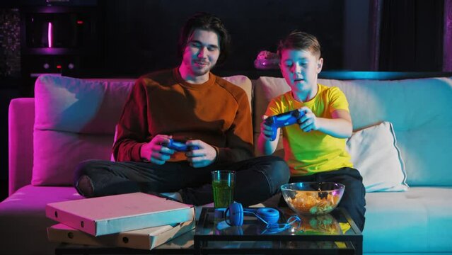 The older brother teaches the younger one how to play the video game console. Together, they play and have fun. When they win, they give each other high fives. Mid shot. 4k