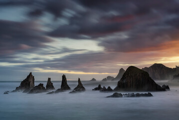 warm sunrise on the beach of Gueirua, Asturias with a dramatic sky and the high tide partially covering the rocks on the shore - obrazy, fototapety, plakaty