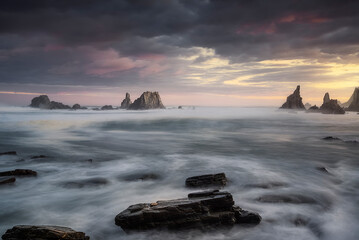 warm sunrise on the beach of Gueirua, Asturias with a dramatic sky and the high tide partially covering the rocks on the shore